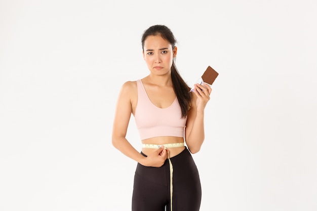 Concepto de deporte, bienestar y estilo de vida activo. Chica asiática triste decepcionada midiendo la cintura con cinta métrica y enfurruñado porque no puede comer barra de chocolate mientras pierde peso con la dieta.