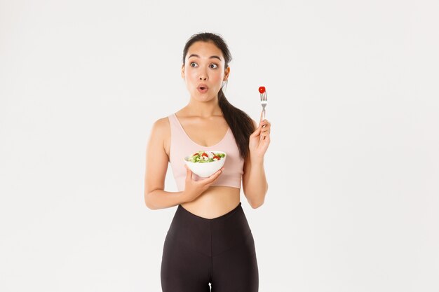 Concepto de deporte, bienestar y estilo de vida activo. Divertida y sorprendida chica de fitness asiática delgada linda en ropa deportiva mirando hacia la izquierda en su logotipo o anuncio mientras come ensalada, fondo blanco.