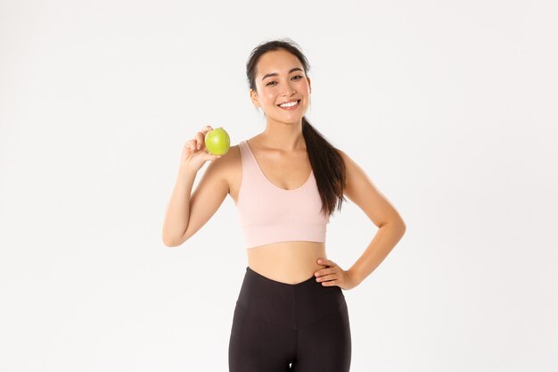 Concepto de deporte, bienestar y estilo de vida activo. Entrenador de fitness femenino asiático atractivo descarado, entrenador de niña en consejos de ropa deportiva comer alimentos saludables después del entrenamiento y entrenamiento, de pie con la manzana.