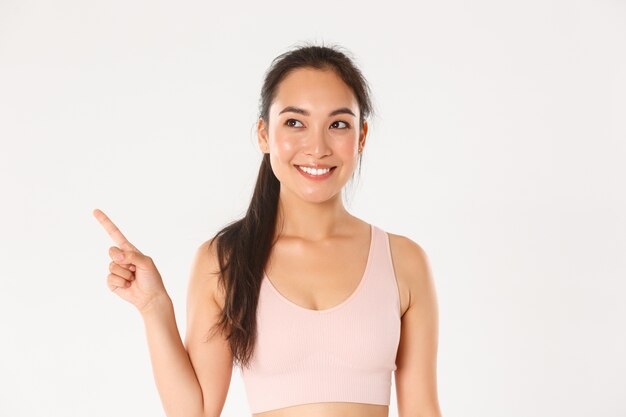 Concepto de deporte, bienestar y estilo de vida activo. Primer plano de una atractiva chica asiática fitness sonriente, atleta femenina recogiendo equipo nuevo en la tienda, apuntando y mirando hacia la esquina superior izquierda complacida.