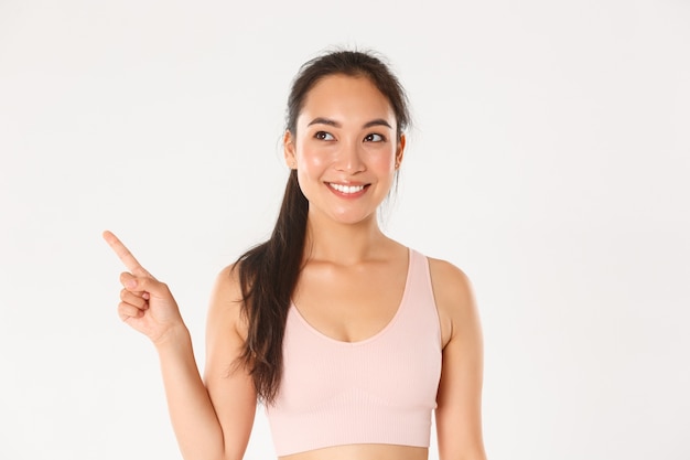 Concepto de deporte, bienestar y estilo de vida activo. Primer plano de una atractiva chica asiática fitness sonriente, atleta femenina recogiendo equipo nuevo en la tienda, apuntando y mirando hacia la esquina superior izquierda complacida.