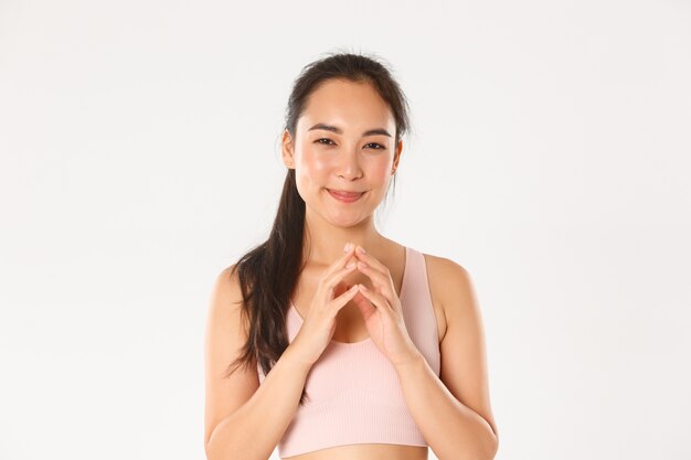 Concepto de deporte, bienestar y estilo de vida activo. Retrato de intrigante pensativa chica asiática fitness, deportista que tiene un plan tortuoso, sonriendo astucia y dedos de aguja, fondo blanco de pie.