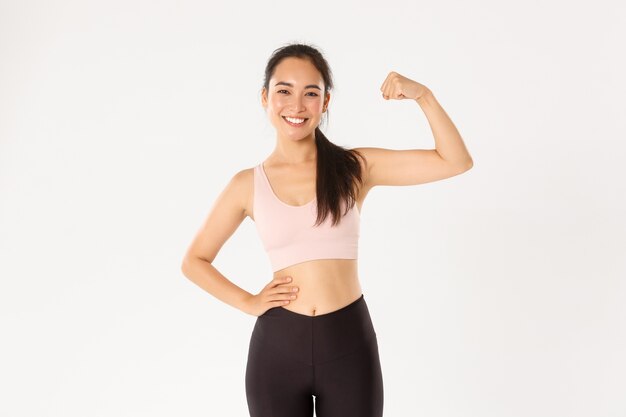 Concepto de deporte, bienestar y estilo de vida activo. Retrato de sonriente chica asiática delgada y fuerte fitness, entrenador personal de entrenamiento mostrando músculos, flexionando bíceps y mirada orgullosa, fondo blanco.