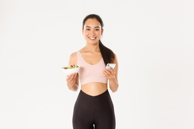 Concepto de deporte, bienestar y estilo de vida activo. Sonriente chica delgada asiática fitness en ropa deportiva, sosteniendo ensalada y teléfono móvil, usando la aplicación de recordatorio de comer, aplicación de control de dieta, control de calorías.