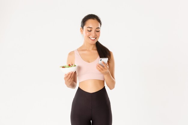 Concepto de deporte, bienestar y estilo de vida activo. Sonriente linda chica asiática que usa la aplicación de dieta, la aplicación de seguimiento de calorías en el teléfono móvil, comuníquese con el entrenador para informar sobre el consumo de alimentos, sostenga la ensalada