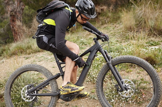 Concepto de deporte, estilo de vida activo, fitness, extremo y adrenalina. Tiro al aire libre del apuesto motorista en equipo de protección presionando los botones en el panel de control en su pedelec, cambiando el modo de velocidad