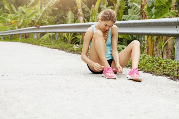 Concepto de deporte y estilo de vida saludable. Deportiva joven sentada en la carretera atando sus zapatillas rosa durante el ejercicio de jogging al aire libre.