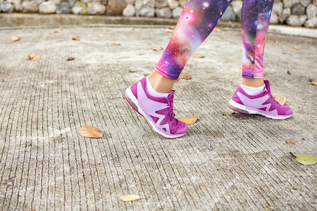 Concepto de deporte, fitness y estilo de vida saludable. Ciérrese encima del tiro de pies femeninos en zapatillas de deporte púrpuras en el pavimento. Deportista en el espacio con leggings estampados y elegantes zapatillas para correr en la carretera en el parque