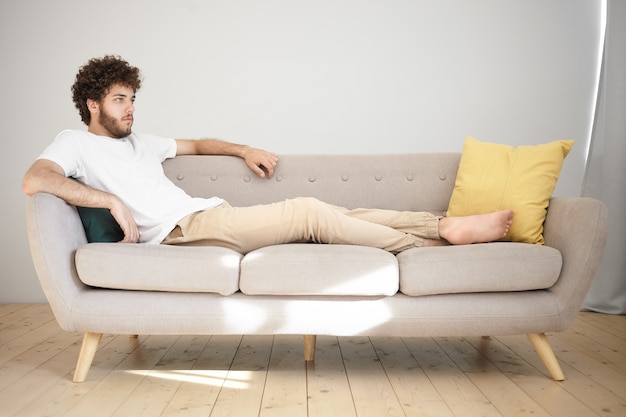 Concepto de descanso, relajación y ocio. Atractivo joven con barba y cabello voluminoso acostado cómodamente en un sofá gris en la sala de estar y viendo la televisión, disfrutando de un partido de fútbol o una serie