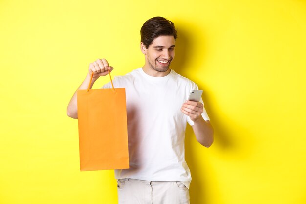 Concepto de descuentos, banca online y cashback. Chico feliz mostrando el bolso de compras y mirando satisfecho en la pantalla del móvil, fondo amarillo.