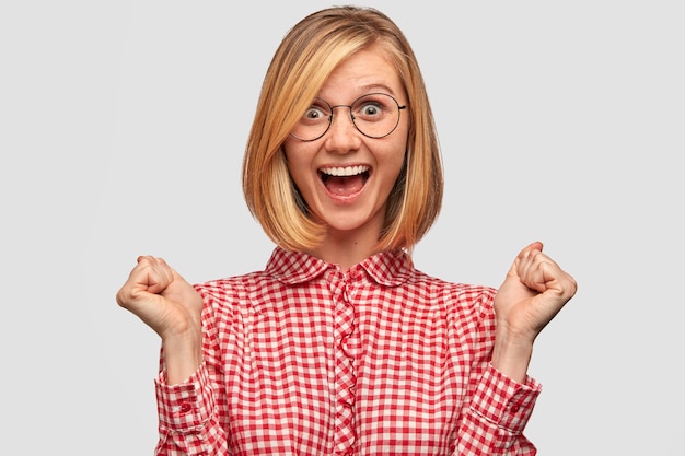 Concepto de determinación y logro de personas. Mujer alegre con saludos de peinado de moda, celebra el éxito, aprieta los puños y exalta sí, vestida con una elegante camisa de moda, aislada en la pared blanca