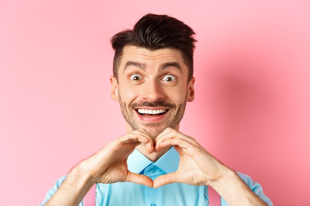 Concepto de día de san valentín cerca de chico romántico que parece feliz sonriendo y mostrando gesto de corazón stan ...