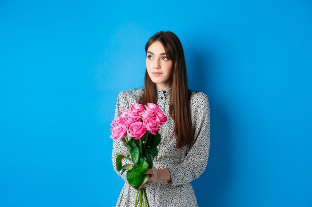 Concepto de día de San Valentín. Chica muy romántica mirando soñadora en el espacio vacío, sosteniendo un ramo de rosas rosadas en la fecha, de pie solo sobre fondo azul.