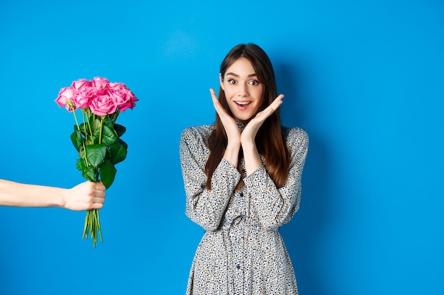 Concepto de día de San Valentín. Mujer joven emocionada y feliz mirando asombrado a la cámara mientras la mano extiende la mano con un ramo de flores, recibiendo un regalo romántico, fondo azul