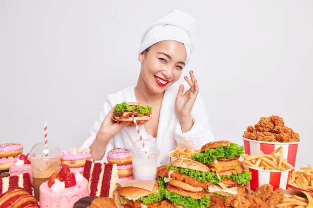 Concepto de dieta poco saludable. La mujer asiática joven positiva con una piel sana sonríe ampliamente sostiene hamburguesa come comida rápida