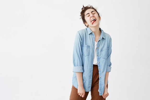 Concepto de diversión, belleza y estilo de vida. Foto de melancólica positiva chica de pelo oscuro sacando la lengua vestida con camisa vaquera sobre camiseta blanca divirtiéndose en el interior