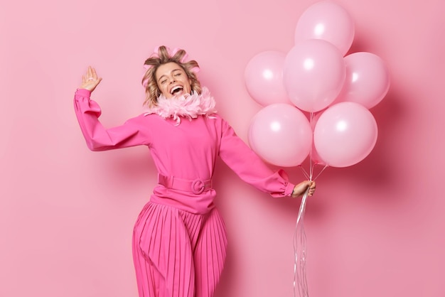 Concepto de diversión y celebración de la gente La mujer alegre y despreocupada usa un vestido largo aplica rulos para el cabello celebra el cumpleaños sostiene un montón de globos de helio aislados sobre fondo rosa baila con alegría