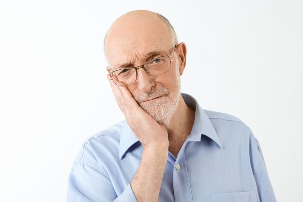 Concepto de edad y personas maduras. Disparo aislado de un pensionista masculino europeo molesto con la cabeza calva y barba gruesa tocando la mejilla, que sufre de dolor de muelas intolerable, con una mirada miserable y dolorosa