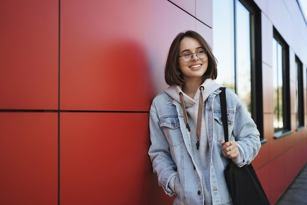 Concepto de educación, estilo de vida y generación joven. Retrato al aire libre de una niña feliz en su camino a casa después de las clases, mirando de reojo soñadora y feliz sonriendo, sosteniendo la bolsa de asas, edificio rojo magro.