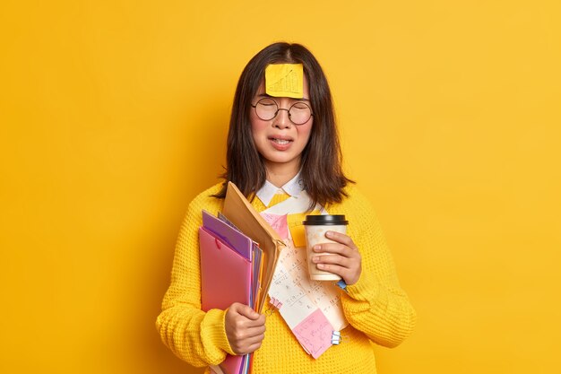 Concepto de educación y personas. Estudiante asiática molesta se siente cansada de la preparación de exámenes, bebidas, café para llevar, calcomanías de notas, tiene mal humor, no puede recordar, todo contiene carpetas con papeles