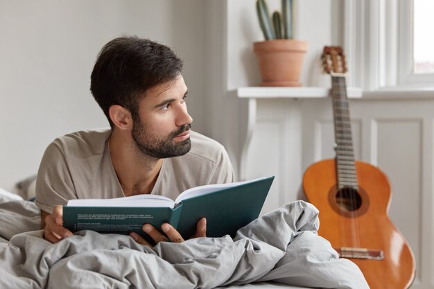 Concepto de educación y preparación de exámenes. Joven sin afeitar yace en la cama, sostiene el libro