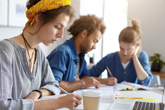 Concepto de educación, universidad y personas. Equipo de estudiantes amigables que trabajan juntos mirando con expresiones serias en sus cuadernos escribiendo con lápices usando una computadora portátil para estudiar