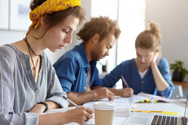 Concepto de educación, universidad y personas. Equipo de estudiantes amigables que trabajan juntos mirando con expresiones serias en sus cuadernos escribiendo con lápices usando una computadora portátil para estudiar