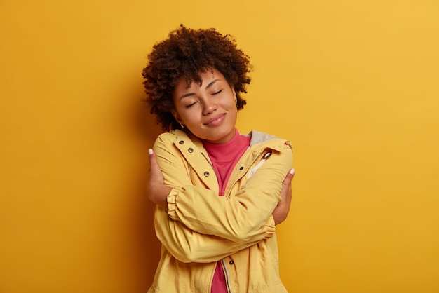 Concepto de egoísmo y amor propio. Retrato de modelo femenina rizada de piel oscura complacida se abraza a sí misma, cruza las manos sobre el cuerpo, mantiene los ojos cerrados, usa chaqueta, posa contra la pared amarilla