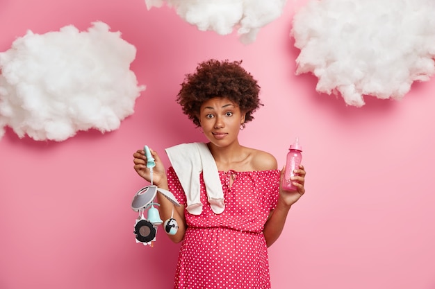 Foto gratuita concepto de embarazo y maternidad. confundida mujer embarazada rizada sostiene cosas de bebé para el feto, se prepara para tener un hijo, posa contra la pared rosa con nubes. futura madre con barriga