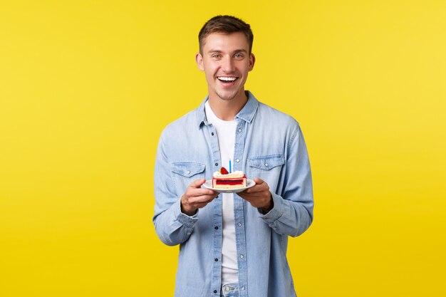 Concepto de emociones de celebración, vacaciones y personas. Alegre joven guapo con fiesta b-day, sosteniendo pastel de cumpleaños con vela y sonriendo, pidiendo deseo sobre fondo amarillo.