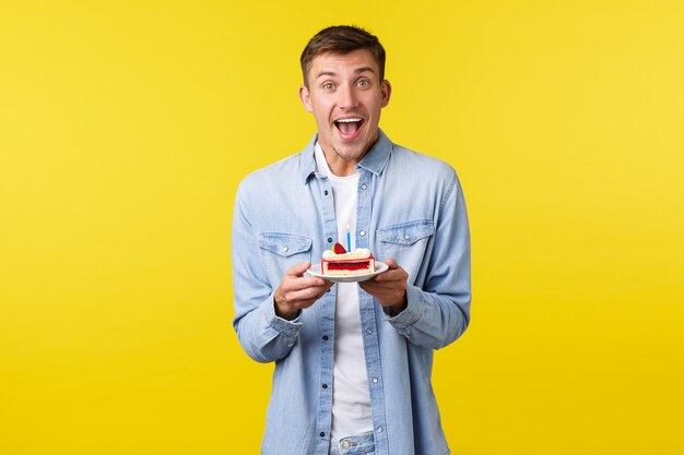 Concepto de emociones de celebración, vacaciones y personas. Hombre feliz entusiasta que sostiene la placa con la torta de cumpleaños, celebrando el b-day, haciendo deseo y velas encendidas, sonriendo emocionado, fondo amarillo.