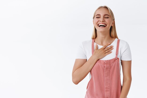 Concepto de emociones, estilo de vida y bienestar. Encantadora mujer rubia caucásica en ropa casual de moda, tocando el pecho mientras se ríe a carcajadas de una broma divertida, cierra los ojos sonriendo ampliamente sintiendo felicidad