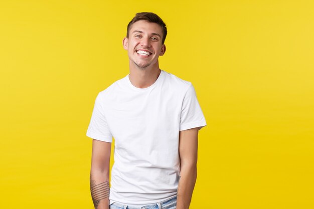 Concepto de emociones de estilo de vida, verano y personas. Chico caucásico carismático guapo en camiseta blanca casual sonriendo ampliamente con una sonrisa blanca perfecta, de pie alegre fondo amarillo.
