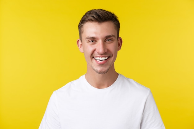 Concepto de emociones de estilo de vida, verano y personas. Modelo masculino guapo entusiasta con sonrisa feliz y dientes blancos, de pie en camiseta casual sobre fondo amarillo satisfecho.