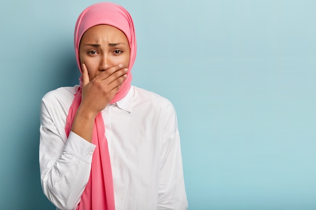 Concepto de emociones humanas negativas. Mujer triste deprimida de raza mixta con hijab llora por las malas noticias, se tapa la boca con la palma, se queja de algo, se para en el interior con un espacio en blanco a un lado para el texto