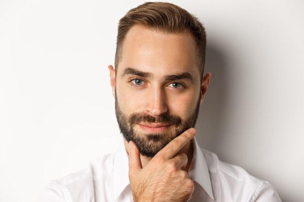 Concepto de emociones y personas. Disparo en la cabeza del hombre guapo pensativo sonriendo satisfecho, tocando la barba y pensando, de pie