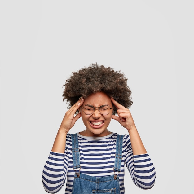 Concepto de emociones y sentimientos negativos. Mujer afroamericana disgustada mantiene las manos en las sienes, vestida de manera informal, posa contra la pared blanca. Mujer de piel oscura tiene dolor de cabeza después del trabajo