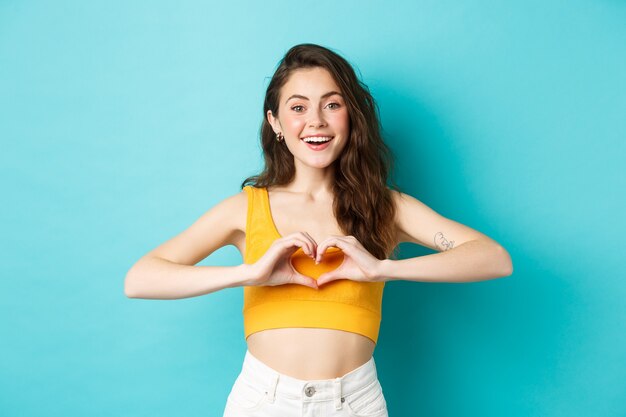 Concepto de emociones y vacaciones de verano. Joven romántica mostrando te amo gesto de corazón y sonriendo, de pie sobre fondo azul.