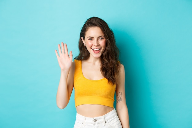 Concepto de emociones y vacaciones de verano. Mujer amable alegre con un cuerpo perfecto, saludando con la mano y saludando, de pie sobre fondo azul.