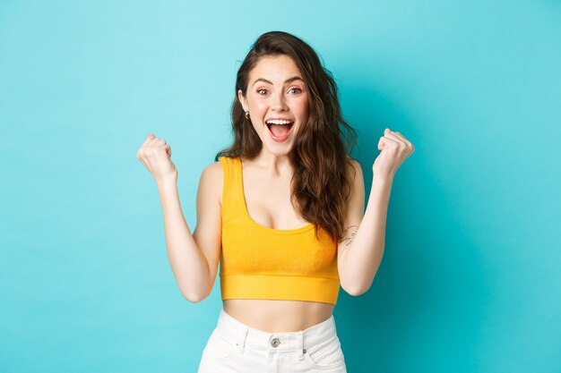 Concepto de emociones y vacaciones de verano. Mujer joven emocionada ganando y gritando de alegría, lograr la meta, celebrando la victoria, haciendo bombas de puño, de pie sobre fondo azul.