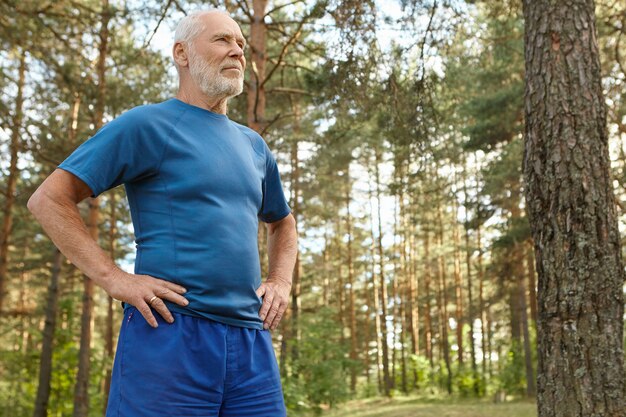 Concepto de energía, salud, bienestar, actividad y deporte. Hombre senior atlético de ajuste concentrado en ropa deportiva manteniendo las manos en la cintura disfrutando de ejercicios físicos en el bosque, de pie entre pinos