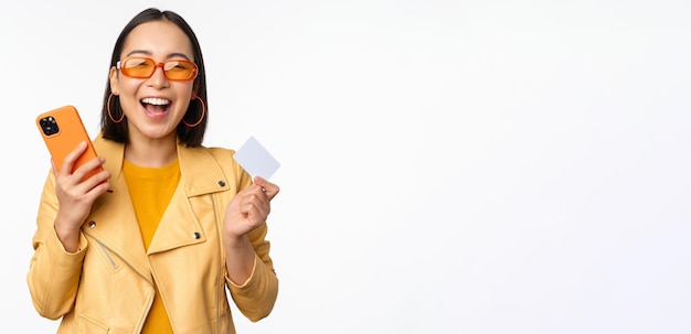 Concepto de entrega y compras en línea Feliz chica coreana en ropa elegante con tarjeta de crédito y teléfono inteligente riendo y sonriendo de pie sobre fondo blanco