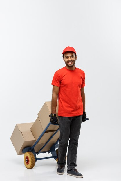 Concepto de entrega - Retrato de apuesto hombre de entrega afroamericana o mensajero empujando camión de mano con pila de cajas. Aislado en el estudio de fondo gris. Espacio De La Copia.