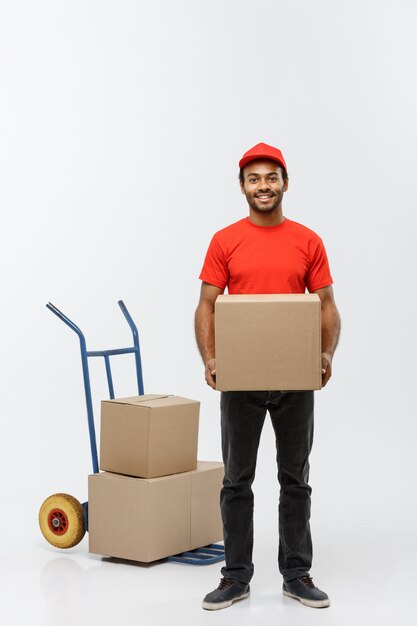 Concepto de entrega - Retrato de apuesto hombre de entrega afroamericana o mensajero empujando camión de mano con pila de cajas. Aislado en el estudio de fondo gris. Espacio De La Copia.