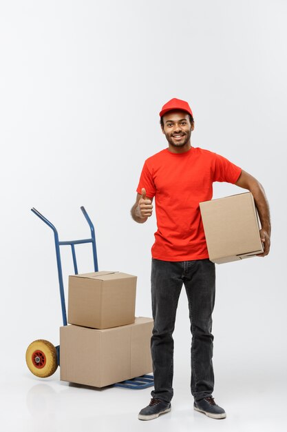 Concepto de entrega - Retrato de apuesto hombre de entrega afroamericana o mensajero empujando camión de mano con pila de cajas. Aislado en el estudio de fondo gris. Espacio De La Copia.