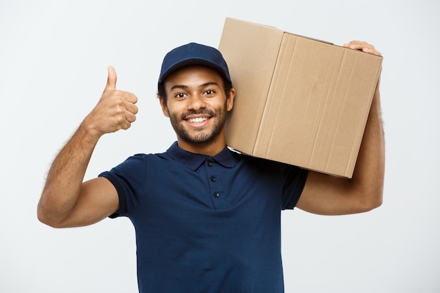 Concepto de entrega - Retrato de feliz africano hombre de entrega de América señalando la mano para presentar un paquete de caja. Aislado en el estudio de fondo gris. Espacio De La Copia.