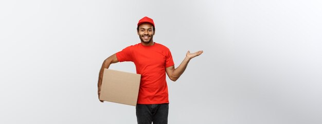 Concepto de entrega Retrato del repartidor afroamericano feliz señalando con la mano para presentar un paquete de caja aislado en el espacio de copia de fondo de estudio gris