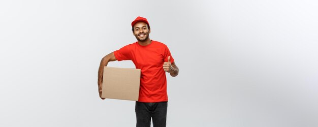 Concepto de entrega Retrato de un repartidor afroamericano feliz sosteniendo un paquete de caja y mostrando golpes aislados en el espacio de copia de fondo de estudio gris