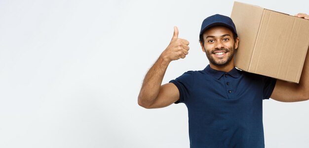 Concepto de entrega Retrato de un repartidor afroamericano feliz sosteniendo un paquete de caja y mostrando golpes aislados en el espacio de copia de fondo de estudio gris