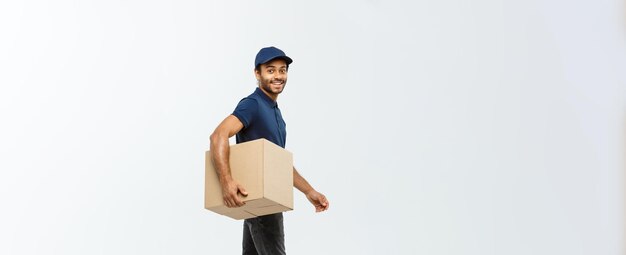 Concepto de entrega Retrato del repartidor afroamericano feliz en tela azul caminando para enviar un paquete de caja al cliente aislado en el espacio de copia de fondo de estudio gris